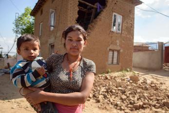 A woman holds a baby in front of a house. the side of the house is missing many of its bricks, which are laying in the yard behind them.