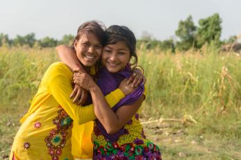 Two teenage girls embracing and smiling in front of a green field in nepal