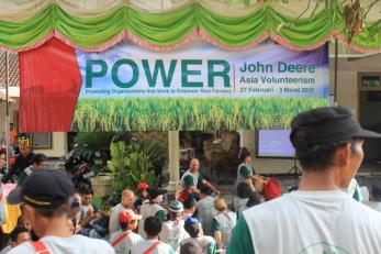 People gathered at an event under a sign that reads: power - promoting organizations that work to empower rice farmers - john deere - asia volunteerism - 27 februari - 3 maret 2017